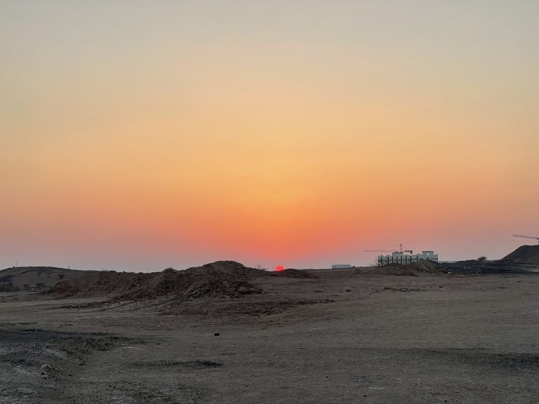 Sunset in the desert. The sun is very low, sand in the foreground a couple of buildings and cranes on the horizon line.