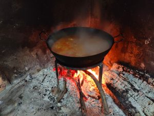 View larger photo: A black pan with liquid and ingredients is placed over a fire with glowing embers. The setting appears to be inside a rustic fireplace with a soot-covered wall in the background.