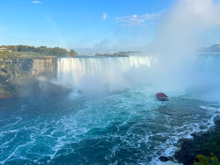 The Maid of the Mist boat steers near to the base of Horseshoe Falls at Niagara Falls on a sunny day, with a faint rainbow toward the left of the image.