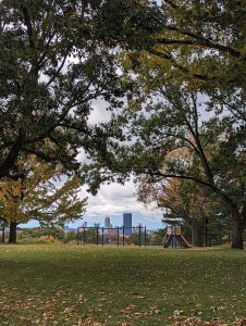 Fall trees with yellow, red and green leaves, with a swing set and the skyline of downtown Pittsburgh in the background 