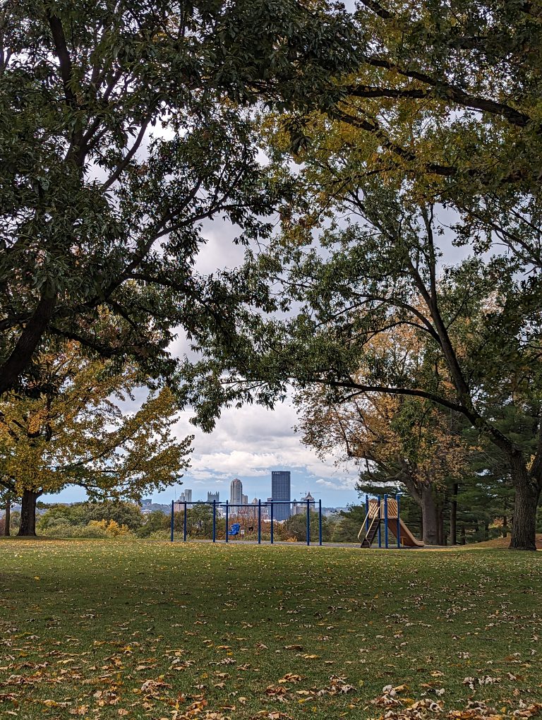 Fall trees with yellow, red and green leaves, with a swing set and the skyline of downtown Pittsburgh in the background