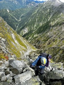 Epic female hiker about to summit Mt. Okuhotaka in the Japanese Northern Alps. A huge drop can be seen behind her as she grabs the steel chain to pull herself upwards. The valley below, with its rocks and greenery, looks majestic.
