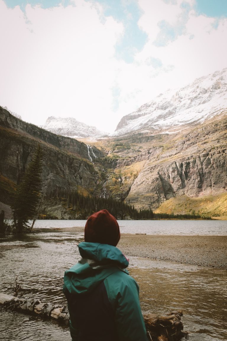 Person in winter hiking gear looking out on a lake with snowy mountains and a waterfall surrounding the lake.