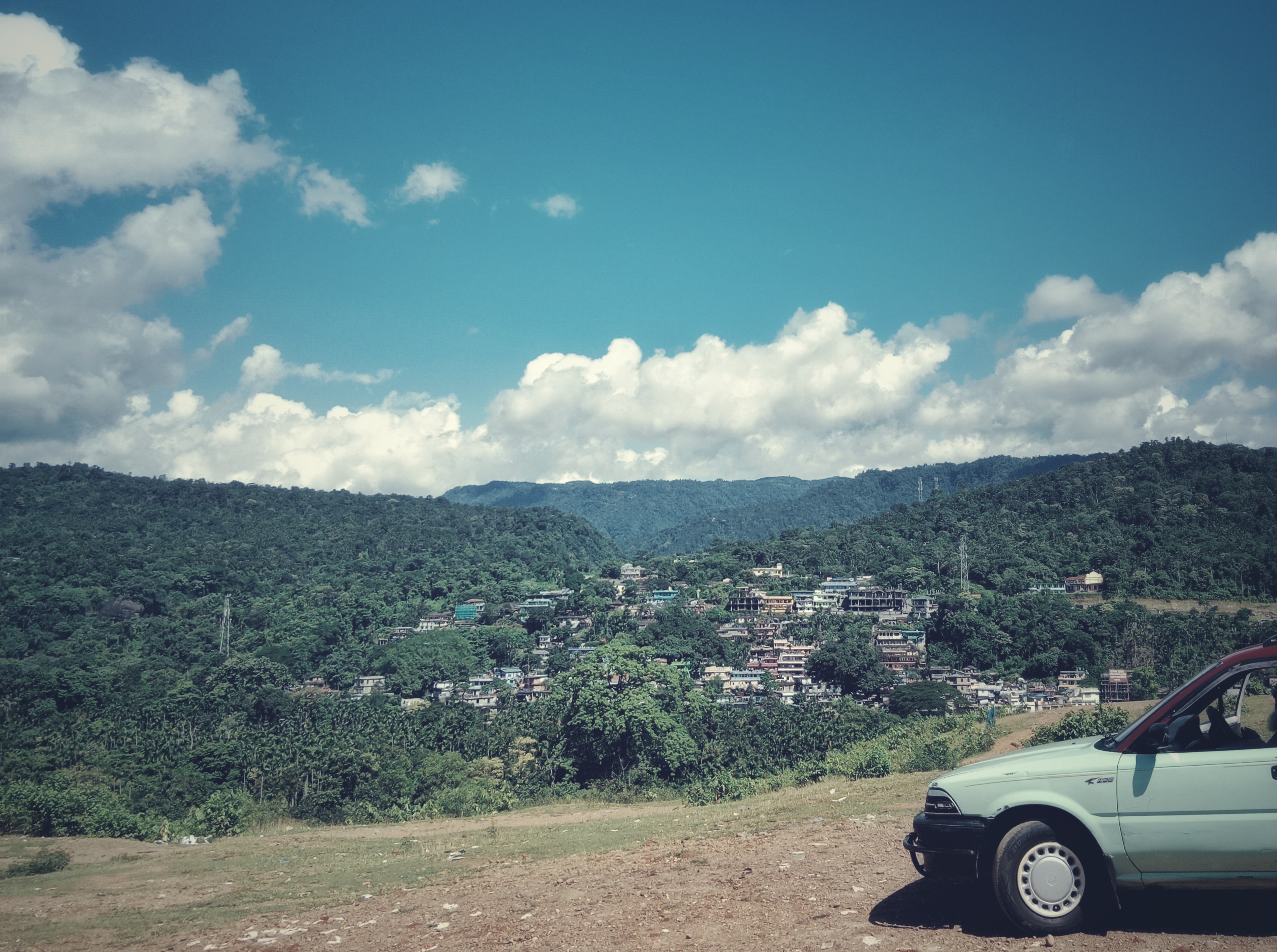 A green car parked on a hilltop overlooking a village.