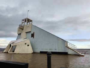 A modernistic structure resembling an old lighthouse stands by the water's edge in Oulu, Finland. The building features a pale blue metallic exterior with a white observation tower topped by a flagpole. A geometric annex, resembling an angular A-frame, is attached. The sky overhead is cloud-streaked.