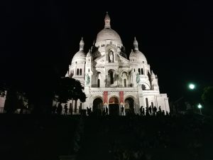 The photo shows the Basilica minor Sacré-Cœur de Montmartre in Paris. A tall building made from white sandstones illuminated in front of a night sky. 
