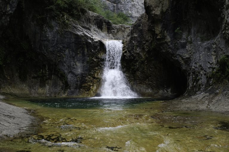 A captivating waterfall cascades into a tranquil pool surrounded by rugged cliffs. The clear, greenish water reveals the rocky bed below. The surrounding cliffs, etched by time, add depth and character to this serene natural setting. The image evokes a sense of peace and untouched beauty.