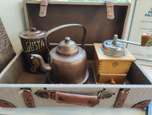 A vintage suitcase holds a copper teapot, a tin with "GUSTA" and a woman's illustration, and a wooden box with a metal grinder on top. The suitcase has brown leather straps and buckles.