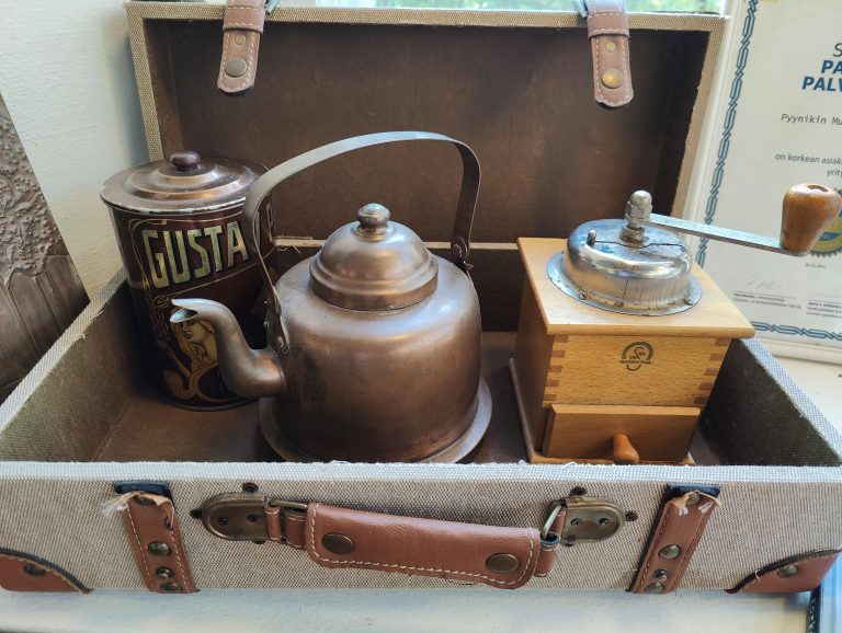 A vintage suitcase holds a copper teapot, a tin with “GUSTA” and a woman’s illustration, and a wooden box with a metal grinder on top. The suitcase has brown leather straps and buckles.