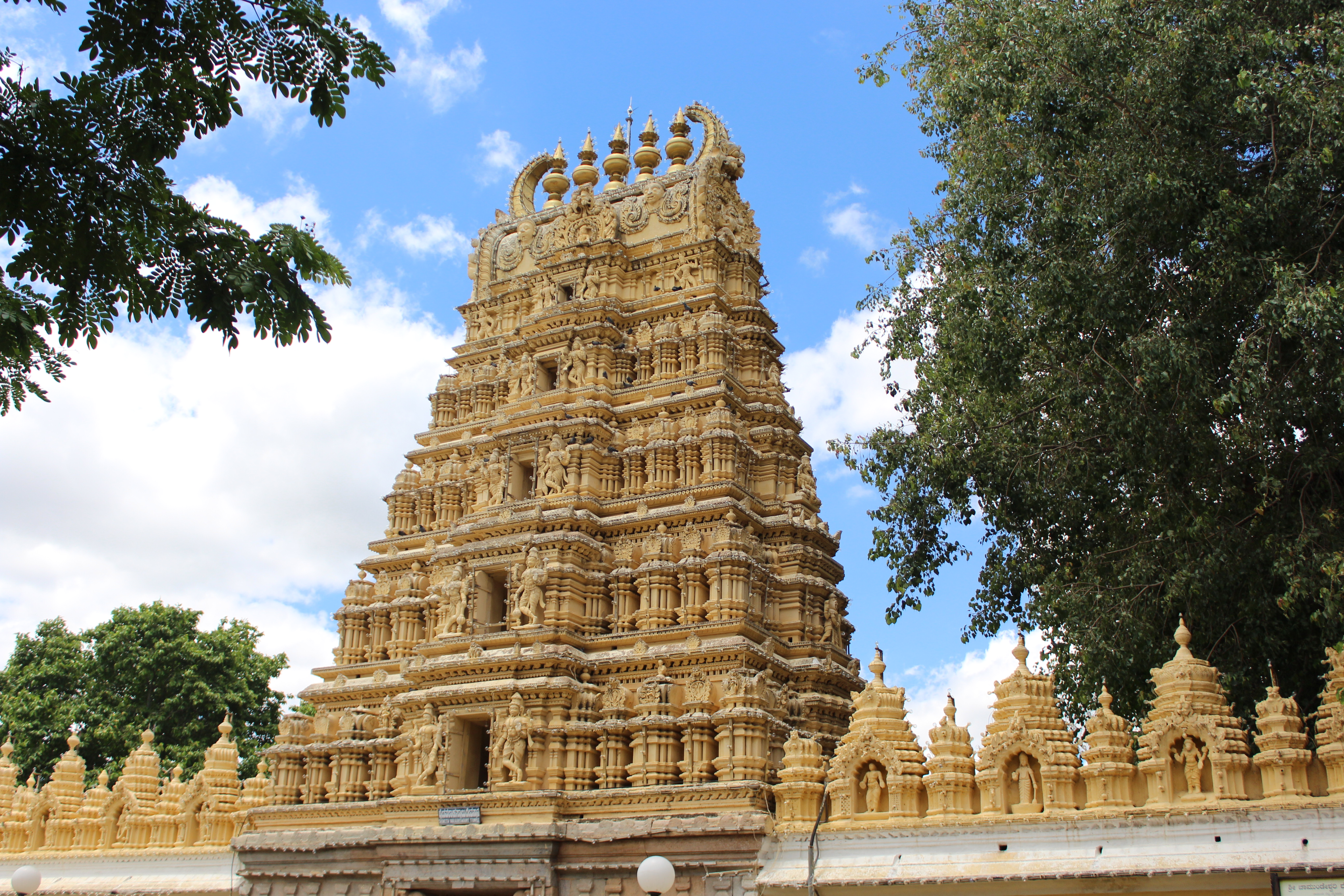 Mysore temple, the temple has a yellow color like sandstone, on the right there is a big green tree, on the left we see the sky on a sunny day.
