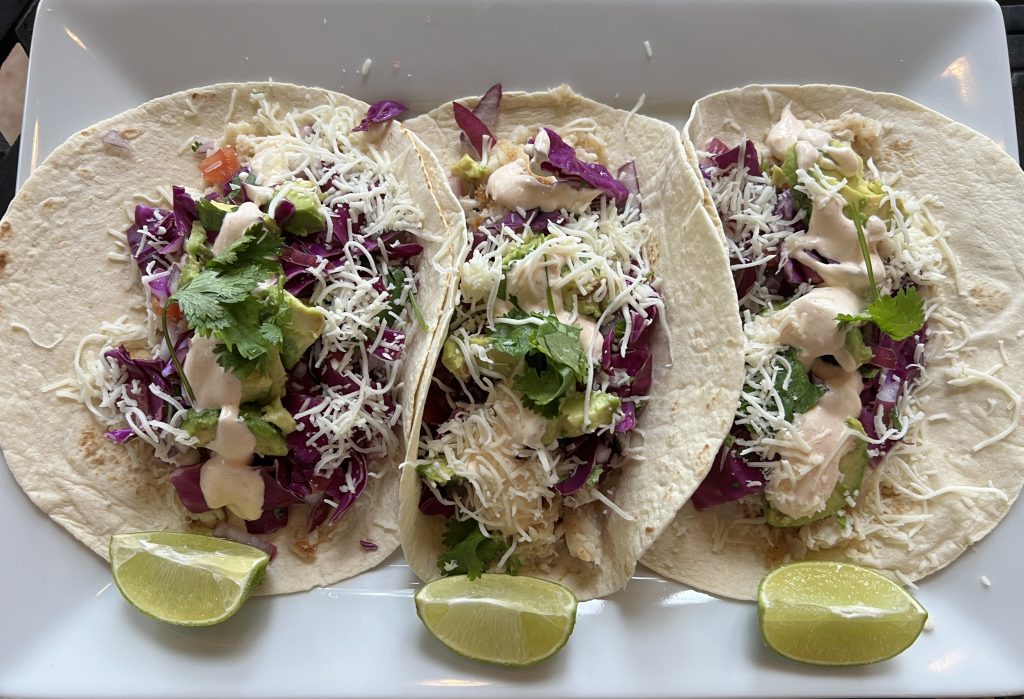 Fish tacos with flaky white fish, shredded cabbage, tomato, sauce, and shredded cheese sit on a plate with three lime wedges.