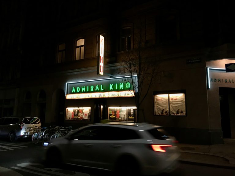 A Viennese old-school cinema facade at night with a car passing in front of it