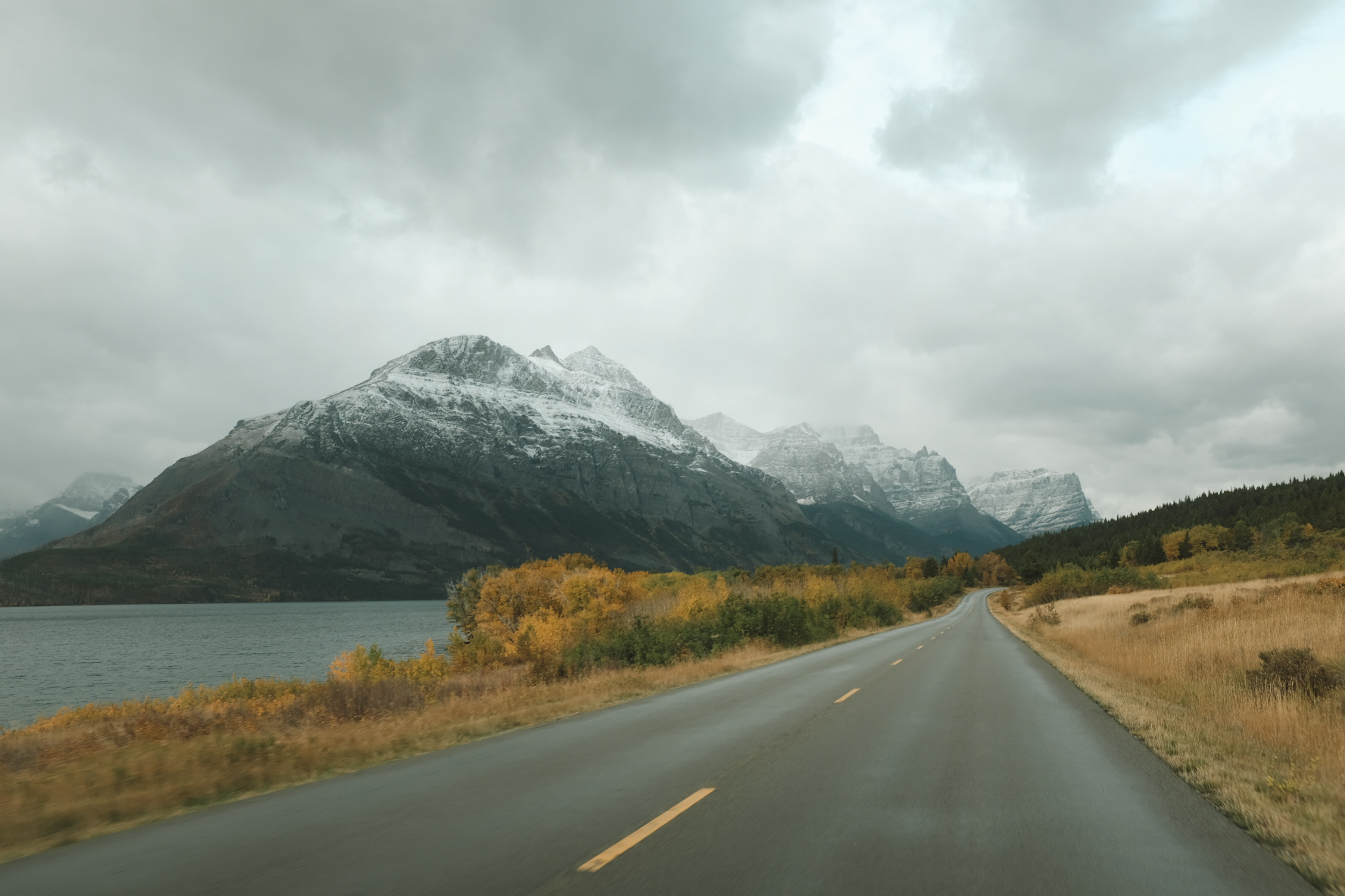 Road curving to the right with snowy mountains in the distance and yellow fall trees. 