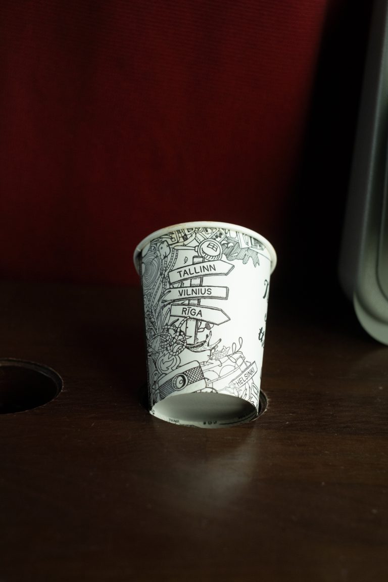 A decorative paper cup with Tallinn, Vilnius, Riga, and Helsinki written on it in the form of a signpost, lying on a wooden surface.