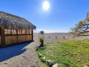 A thatched-roof structure with bamboo walls stands next to a gravel path. There's a tree with fallen branches nearby. In the distance is a sandy beach, a barrier, and the sea under a clear sky with the sun shining.