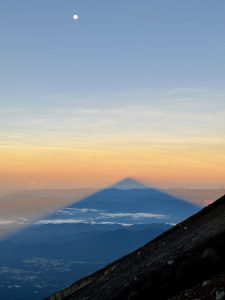 The breathtakingly perfect triangle shape of the shadow of Mt. Fuji at dawn from its highest point: Kengamine Peak. The full moon can be seen hanging from the steel blue sky. The rocky, steep incline contrasts with the blue and yellow hues of the sky. A charming Japanese town is visible down below.
