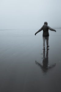 Person walking away on a rainy beach with the arms open to the side and their reflection visible on the shore. 