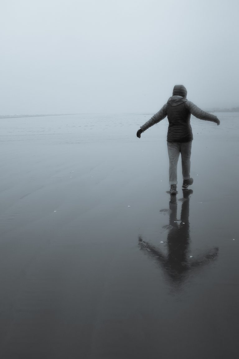 Person walking away on a rainy beach with the arms open to the side and their reflection visible on the shore.