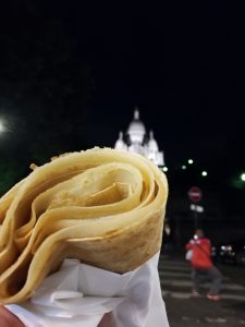A detail shot of a rolled up Crépe in the front and in the back the illuminated building of Sacré-Cœur in the night sky
