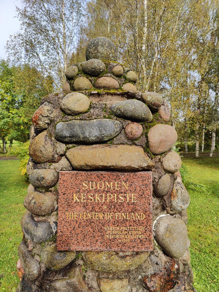 A stone monument composed of various-sized rounded rocks stands against a backdrop of tall trees. Affixed to the monument is a rust-colored plaque inscribed with “SUOMEN KESKIPISTE” and “THE CENTER OF FINLAND.” The plaque also references a date: 1956. Surrounding the monument is a lush green meadow.