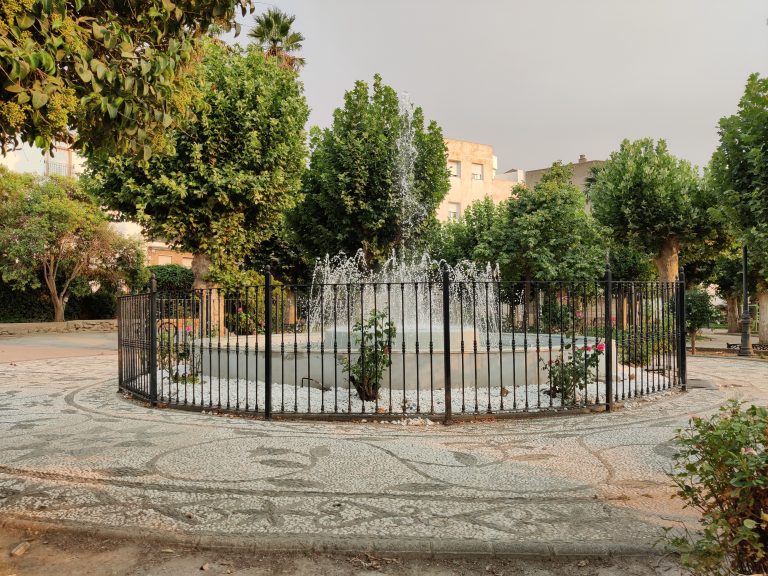 An urban park with a fenced fountain in the center. Surrounding the fountain are various trees, shrubs, and flowering plants. In the foreground, there’s a patterned stone pathway. Buildings are visible in the background under a hazy sky.