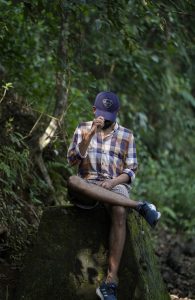 View larger photo: A man sitting on a moss-covered stone. He is tipping his ball cap toward the camera, covering his face. He is wearing a plaid shirt and shorts. Trees are in the background. #WPPhotoFestival