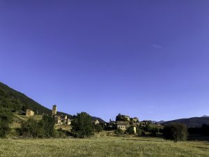 A quaint village nestles against a verdant hillside, its historic stone buildings punctuated by a prominent church tower. The expansive blue sky overhead contrasts with the green landscape. The village exudes an aura of tranquility, representing a bygone era preserved in time amidst nature’s embrace.
