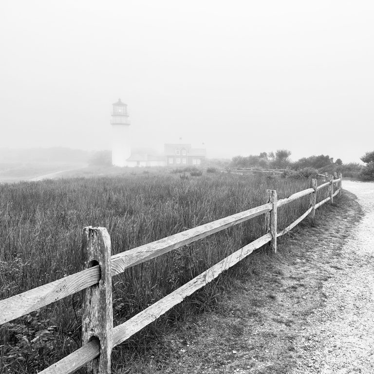 Lighthouse, Cape Cod, USA bordered by the ocean and a field long grass, edged with a ranch style wooden fence, a track runs along side the fence. A monochrome capture on a misty day.