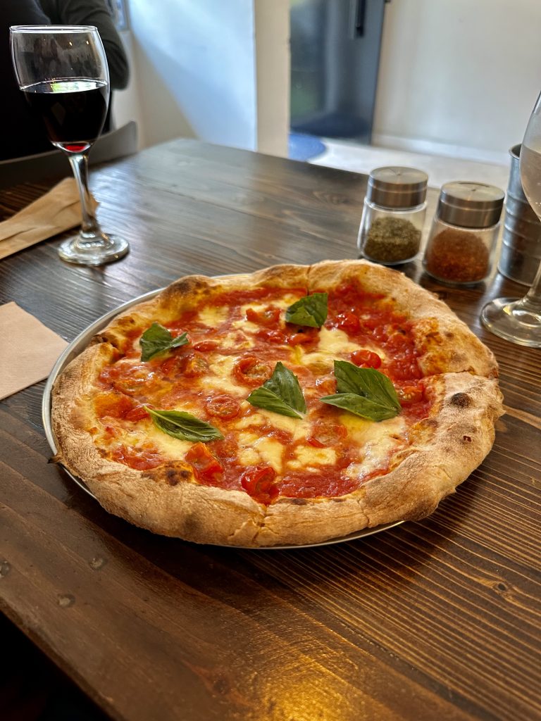Delectable pizza Margherita on top of a wooden table, with a nice toasted rim, fresh basil leaves, and bright tomato sauce spotted with pearl white mozzarella cheese. A tempting glass of red wine sits right next to it. Chili flakes and pepper wait patiently in their containers to become toppings if needed.