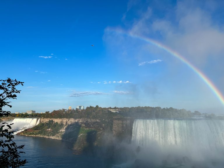 A rainbow rises through the mist of Horseshoe Falls of Niagara Falls. The American falls can be seen further down the gorge to the left. Mist rises like clouds throughout the scene.