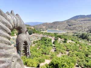 A metal statue of a creature's head with pointed teeth is in the foreground. In the background, there's a landscape with green vegetation, a body of water, and hills under a clear sky.