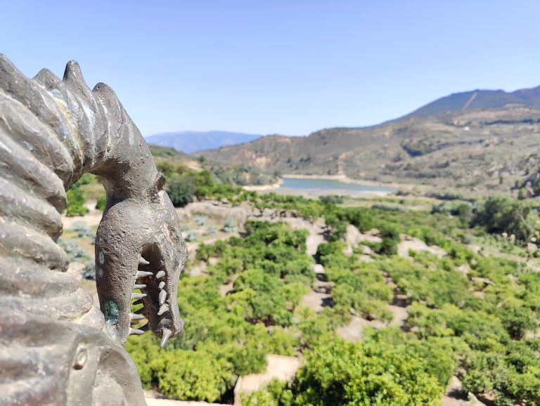 A metal statue of a creature’s head with pointed teeth is in the foreground. In the background, there’s a landscape with green vegetation, a body of water, and hills under a clear sky.