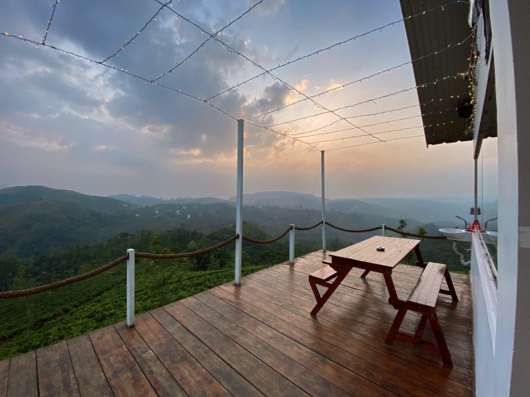 A wooden decked veranda edged with a rope fence, picnic bench and sink with some fairy lights suspended above it. Taken from the top of a hill during the evening. A mountainous lush vista, Vagamon, Kerala.