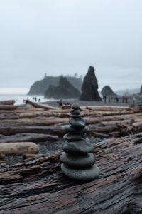 Rock cairns in focus against a backdrop of the rocky, Oregon coast with giant logs. 