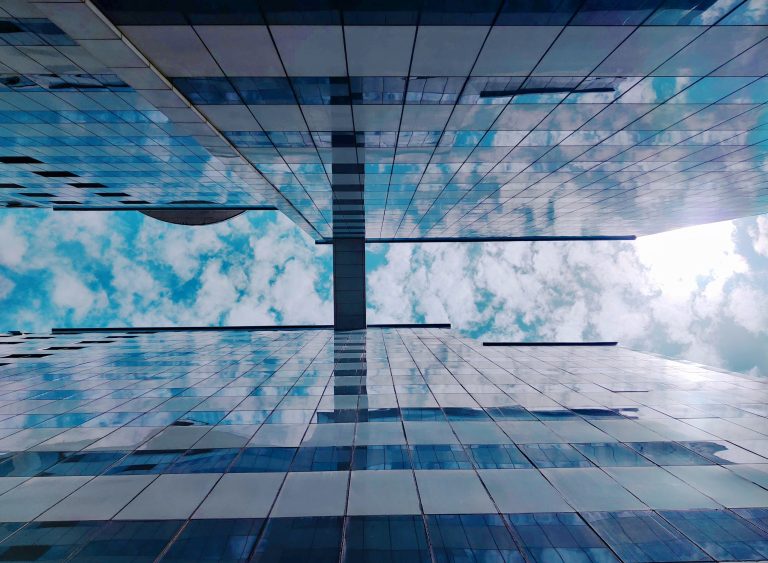 Standing between two modern glass front buildings looking upwards. The blue sky and the clouds are reflecting in the windows. There is some sort of bridge between the two buildings.