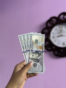 A girl is holding USD in her hand and wall clock in the background.
(Please note rules for using photos of US currency here https://www.bep.gov/currency/currency-image-use)