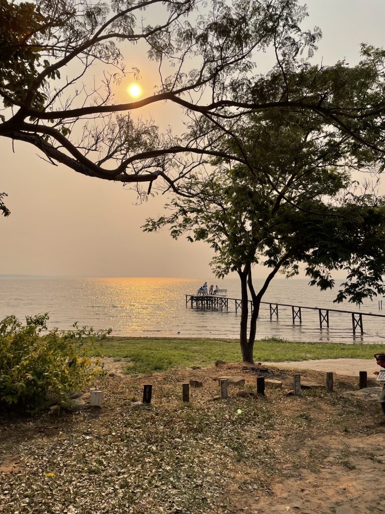 A serene sunset view over a body of water with a silhouetted tree in the foreground and a small pier extending into the water with a few people gathered on it. The sun casts a golden reflection on the water’s surface. “Lago Ypacarai” in paraguay.