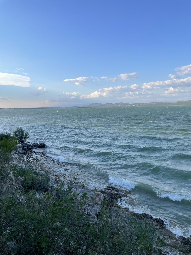 Standing on a cliff to the lake “Vransko jezero, Vrana, croatia” . It is very windy there are big waves for a lake. Far off in the distance on the other side of the lake there are some small mountains. The sky is bright blue, it is a wonderful day.