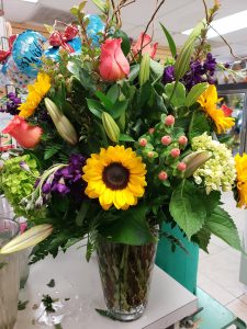 Photo of a large flower arrangement on a flower shop counter. Flowers include roses and sunflowers.
