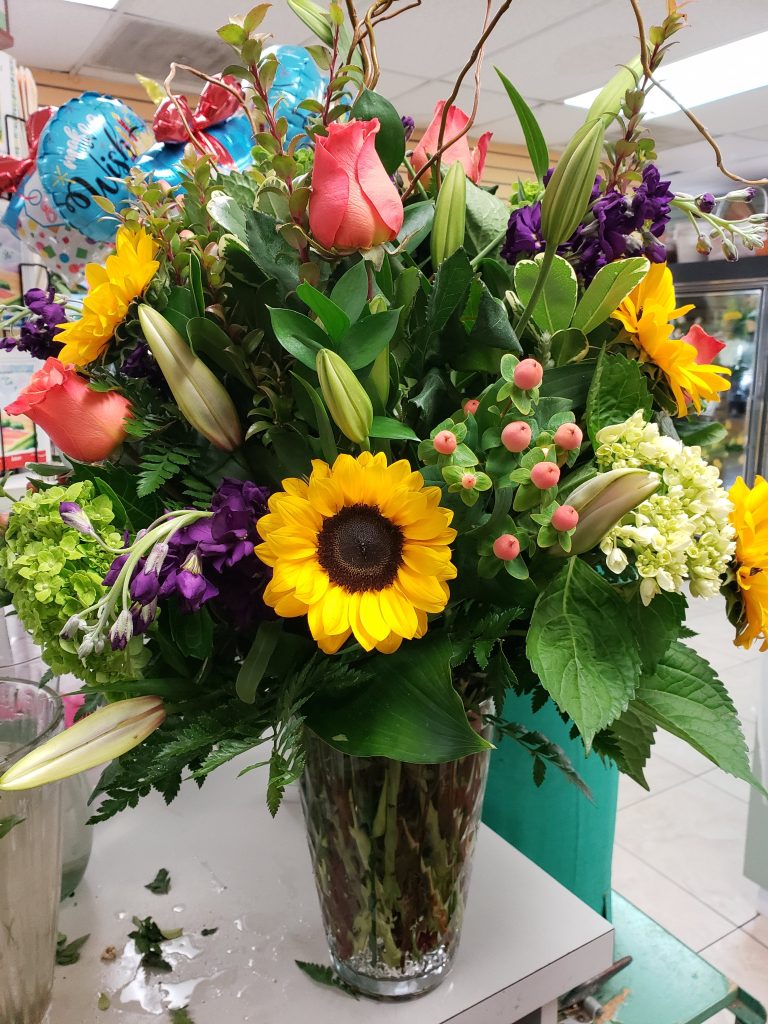 Photo of a large flower arrangement on a flower shop counter. Flowers include roses and sunflowers.