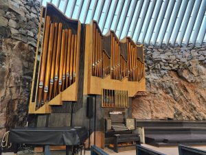 The interior of Temppeliaukio Church in Helsinki, with a large pipe organ against the natural rock wall, under a row of skylight windows.
