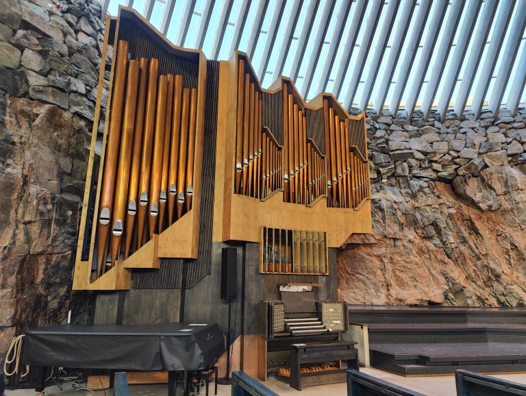 The interior of Temppeliaukio Church in Helsinki, with a large pipe organ against the natural rock wall, under a row of skylight windows.