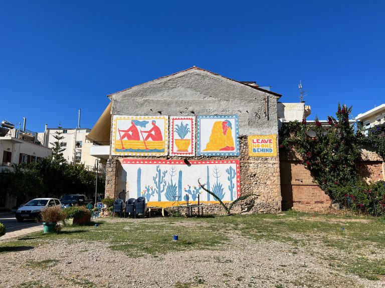 Photo of the wall of a building decorated with yellow, blue and red graffiti murals and the text “Leave No One Behind” taken in Itea, Greece.