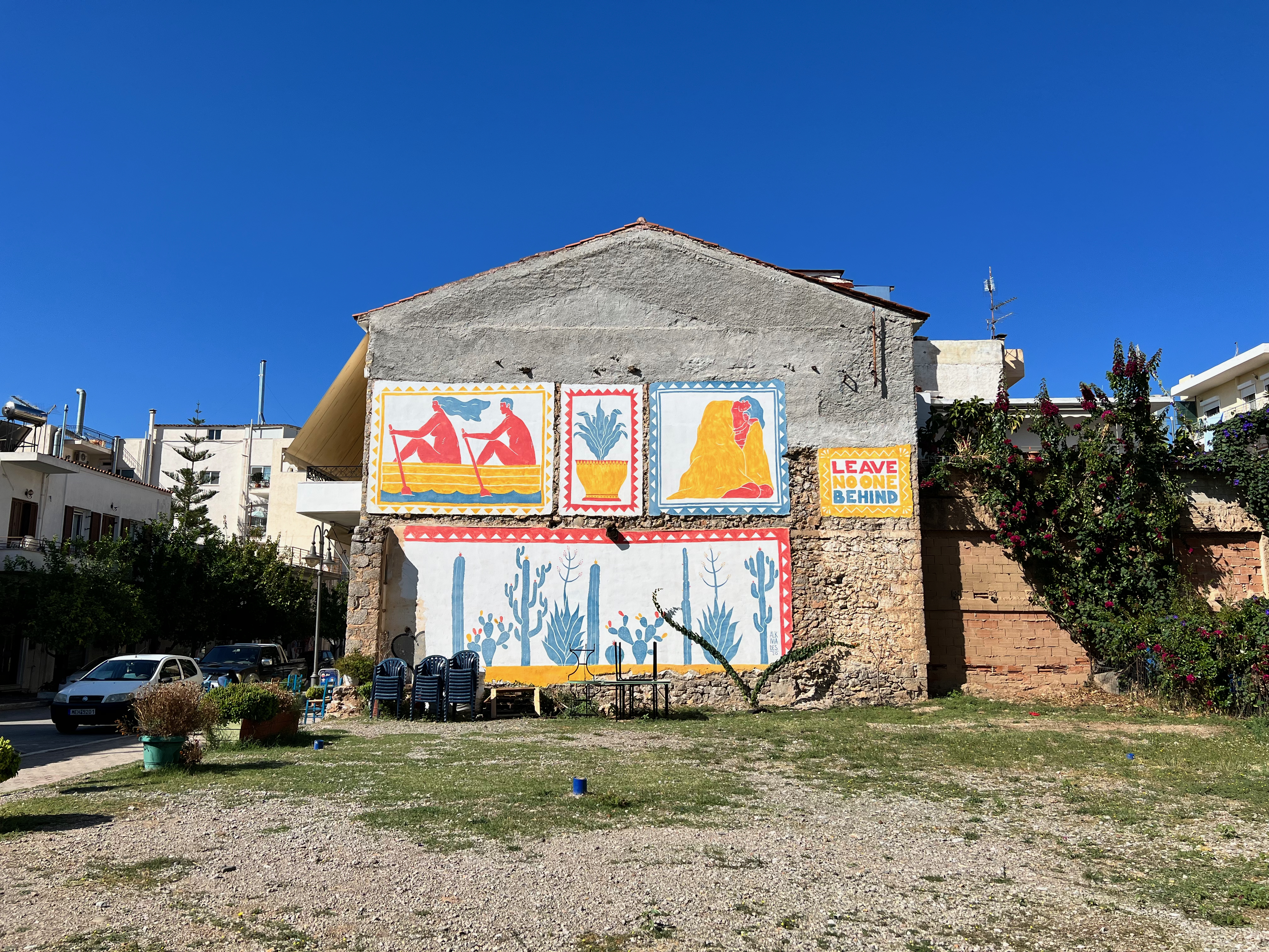 Photo of the wall of a building decorated with yellow, blue and red graffiti murals and the text "Leave No One Behind" taken in Itea, Greece.