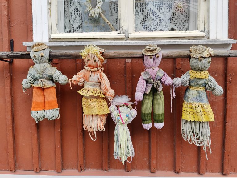 Four handcrafted dolls, made of colorful yarn and fabric, hang against a rust-red wooden wall. Each has distinct outfits, from orange skirts to green pants and lace dresses. Above, a window with white lace curtains displays a decorative sunflower.