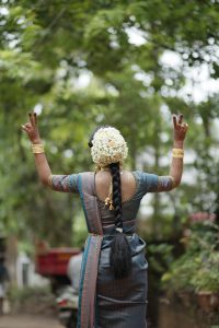 A woman at a wedding walking away from the camera. She has flowers in her hair and henna on her hands. Her hands are up with her fingers in a "V" pose.