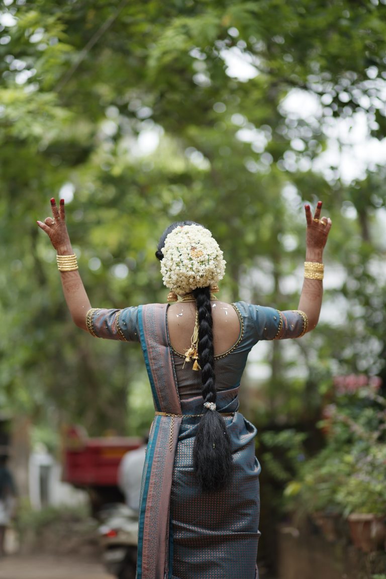 A woman at a wedding walking away from the camera. She has flowers in her hair and henna on her hands. Her hands are up with her fingers in a “V” pose.