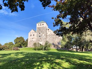 The photo captures Turku Castle in Finland, a majestic medieval structure with a prominent stone tower, set against a serene park with vibrant green grass and trees. The castle, partly in shadow, contrasts with the bright blue sky dotted with wispy clouds.
