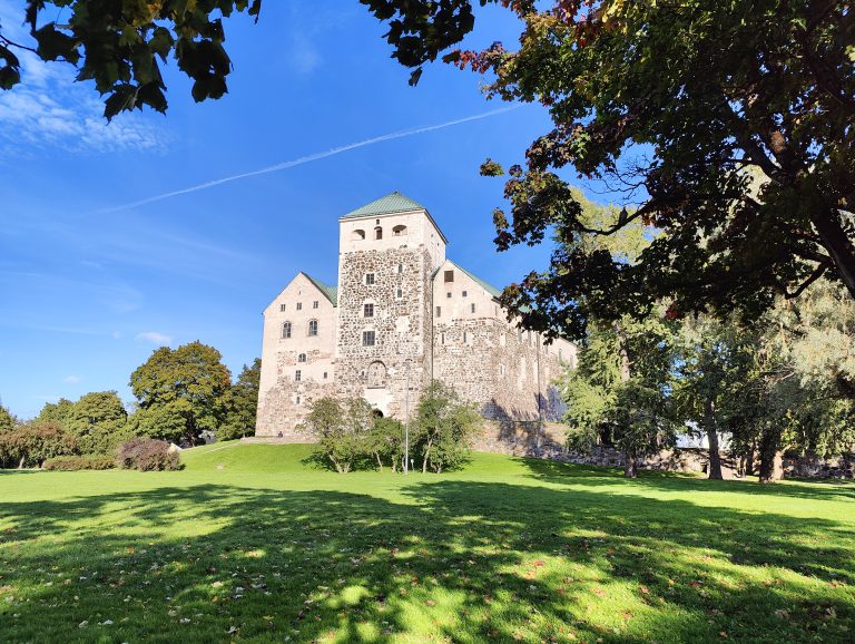 The photo captures Turku Castle in Finland, a majestic medieval structure with a prominent stone tower, set against a serene park with vibrant green grass and trees. The castle, partly in shadow, contrasts with the bright blue sky dotted with wispy clouds.