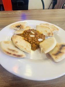 Plate of food including quartered pieces of sharing bread arranged around the edge of the plate, garnished with three nuts. Presented on a table.